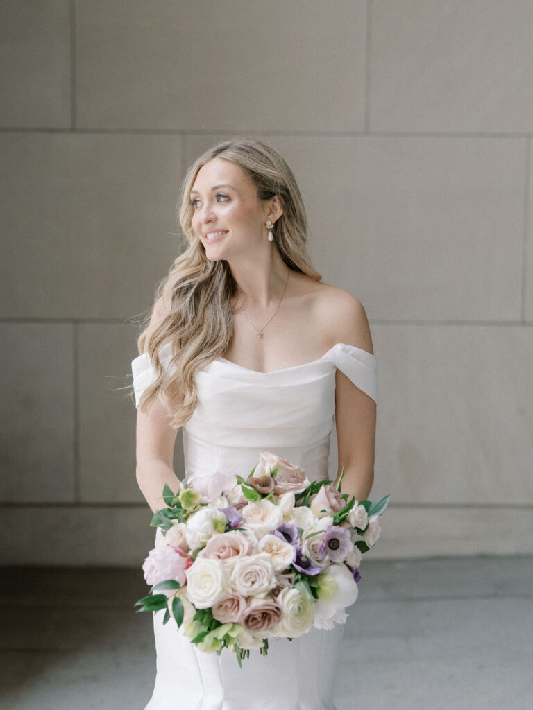 Bride holding a pastel bouquet and smiling off-camera, standing against a neutral stone wall at the Civic Auditorium in downtown Grand Rapids.
