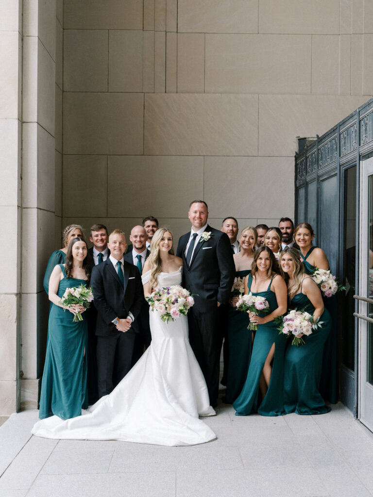 Bride and groom surrounded by their wedding party in emerald green dresses and black suits, standing under the tall limestone arches of the Civic Auditorium.