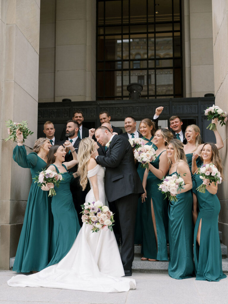 Wedding party cheering with raised bouquets as the bride and groom share a kiss in front of the Civic Auditorium’s detailed stone facade.