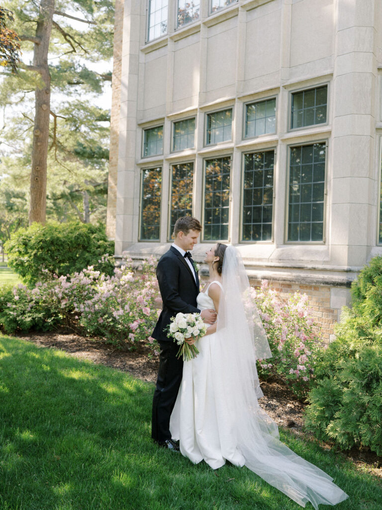 Bride and groom embracing in front of a classic stone building surrounded by spring blooms for their Notre Dame wedding photos