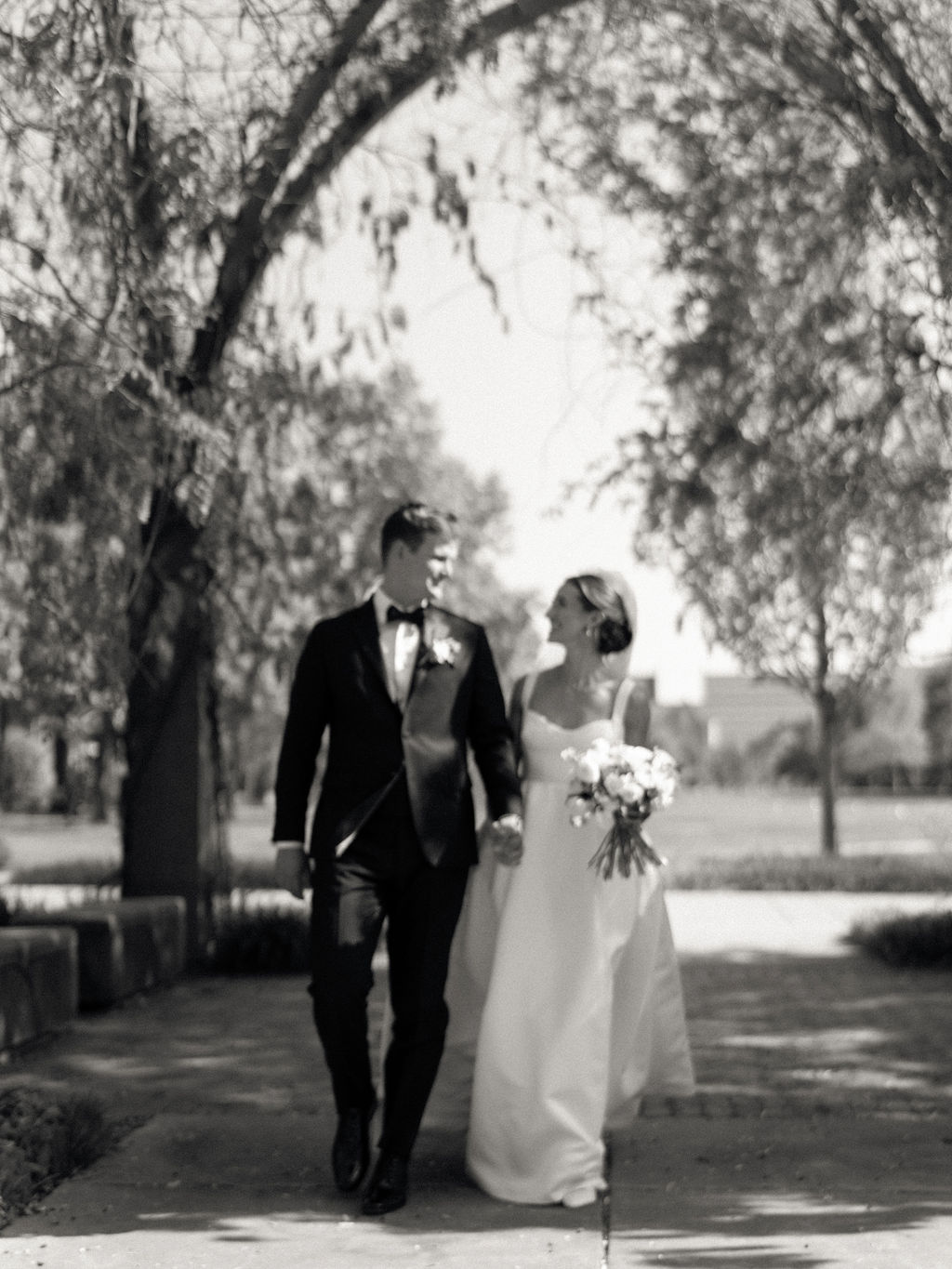 Black and white image of bride and groom walking together under tree-lined shade after their Notre Dame wedding ceremony