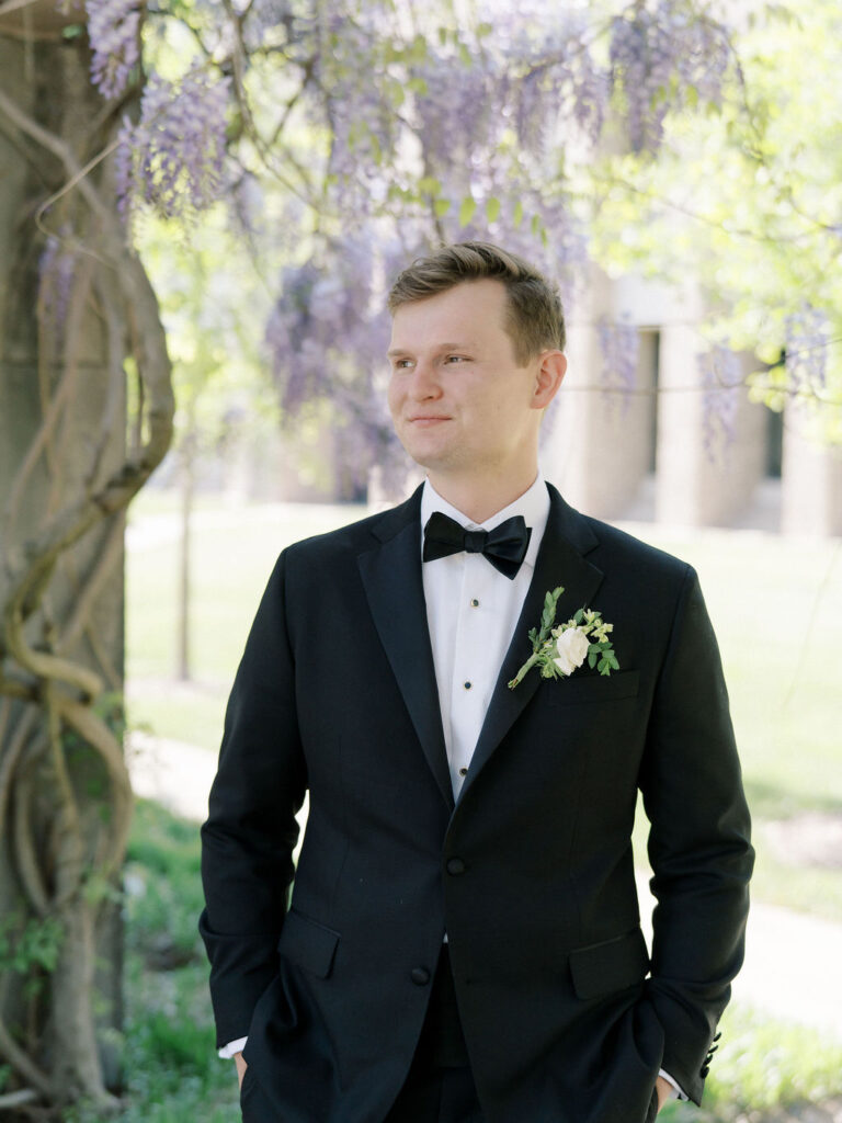 Groom standing beneath wisteria vines, smiling and dressed in a black tux