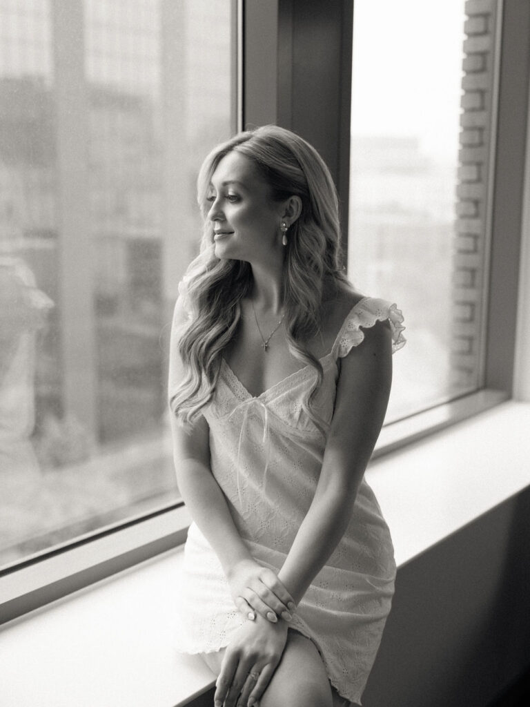 Black and white photo of a bride in her pajamas sitting by her hotel window