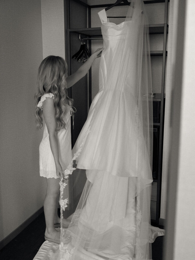 Black and white photo of a bride admiring her hanging wedding dress