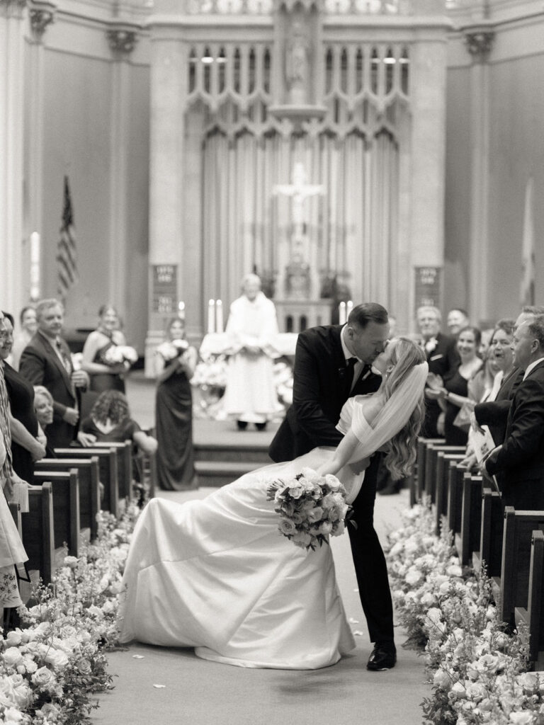 A black-and-white photo of the couple sharing a kiss at the end of the aisle, framed by rows of smiling guests and flowers.