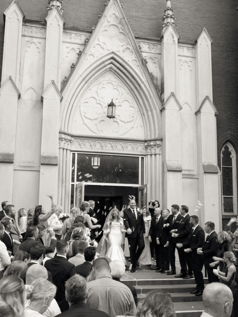 Black-and-white photo of the newlyweds walking out of the church doors while friends and family toss flower petals.