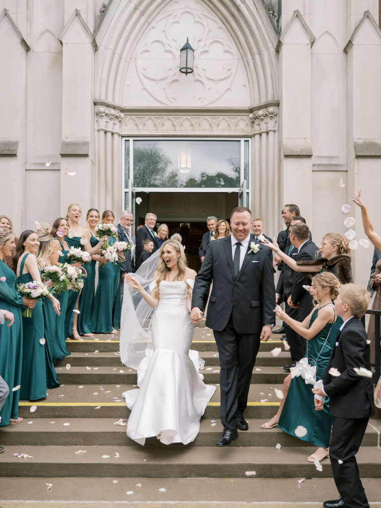 The bride laughs and holds her veil as she and the groom walk through flying petals, full of joy and celebration.
