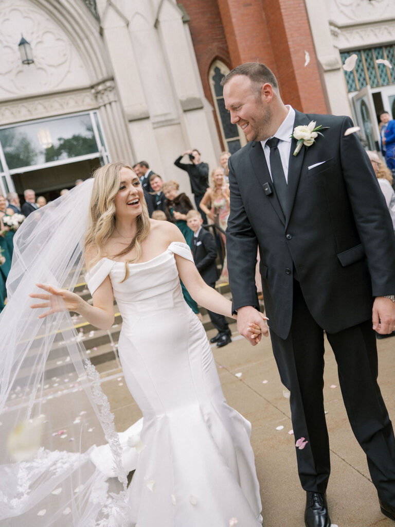 The bride laughs and holds her veil as she and the groom walk through flying petals, full of joy and celebration.