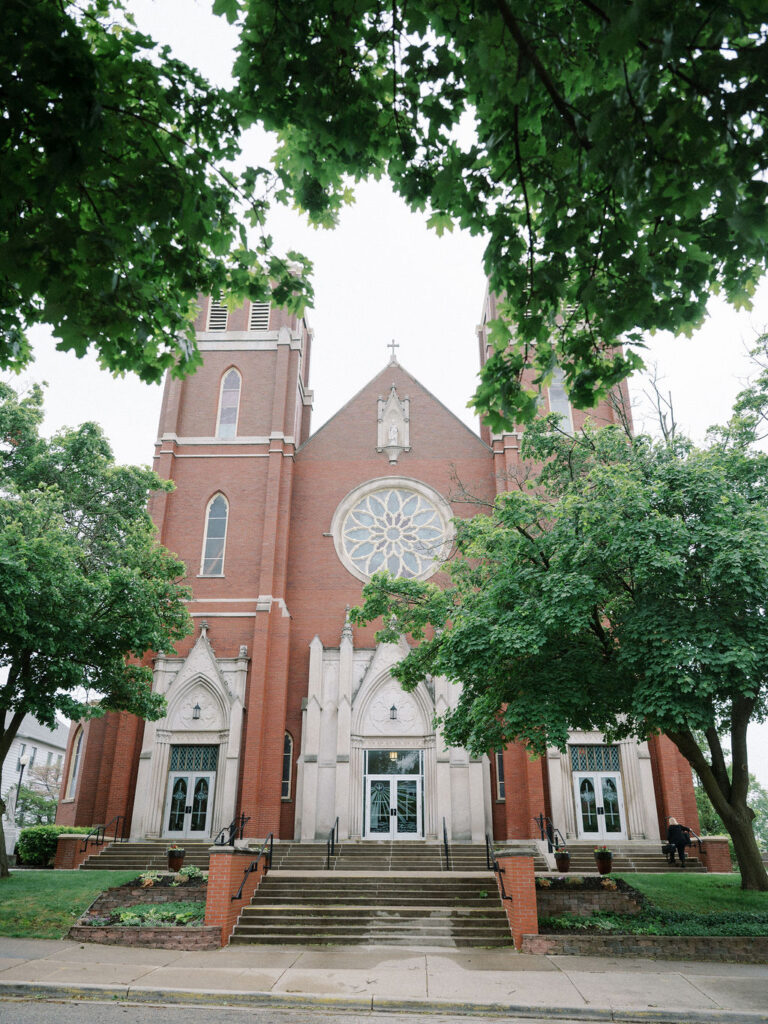 A grand brick church with twin towers and a large rose window, captured from the sidewalk with lush green leaves framing the image