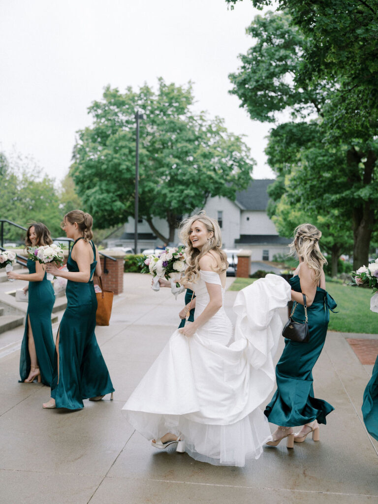 The bride in a fitted white gown smiles as she walks with her bridesmaids, who are wearing deep green satin dresses and holding bouquets.