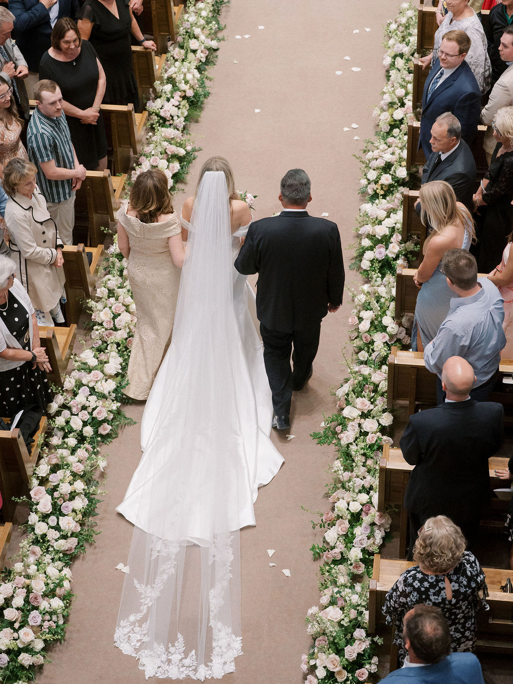 Overhead view of the bride walking through a church aisle lined with blush and white florals, arm in arm with her parents.