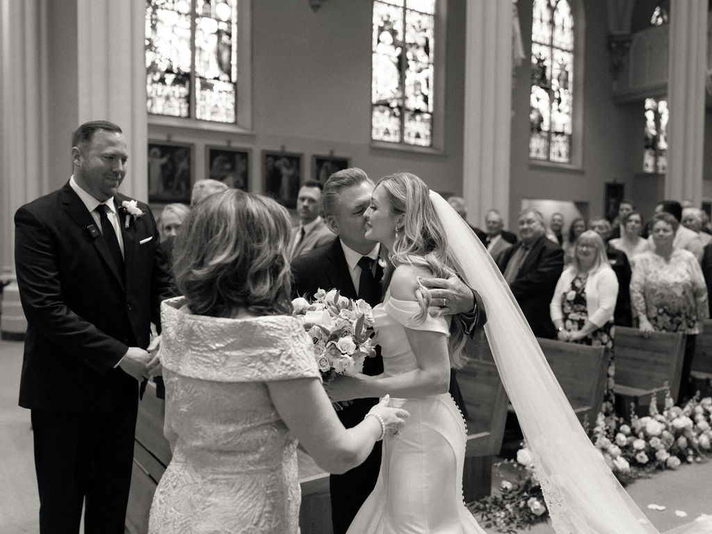 A sweet black-and-white moment as the bride embraces her dad while the groom and guests watch with smiles.