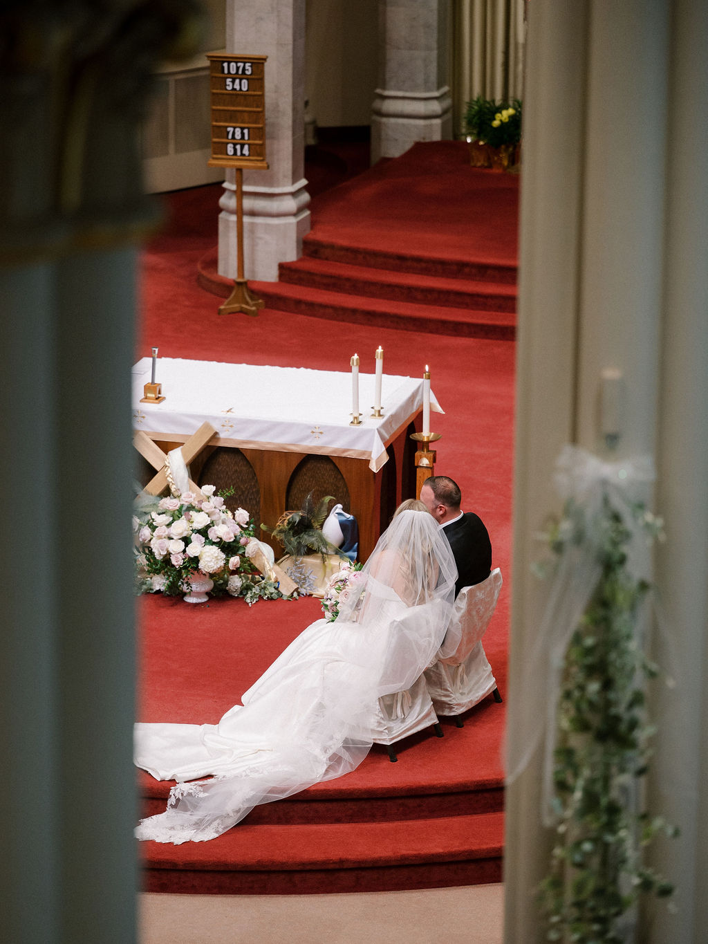 The bride and groom sit side by side with the bride’s veil spilling over the red-carpeted steps, surrounded by floral arrangements.