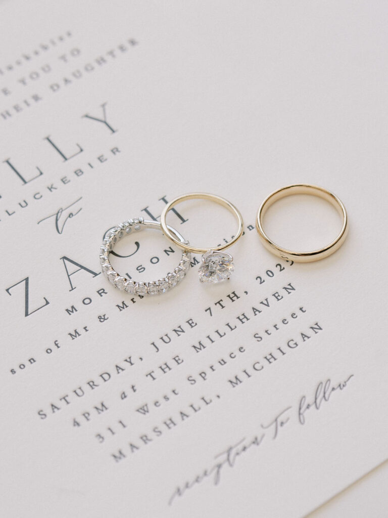 Wedding detail shot of the brides rings and grooms band on top of their wedding invitation