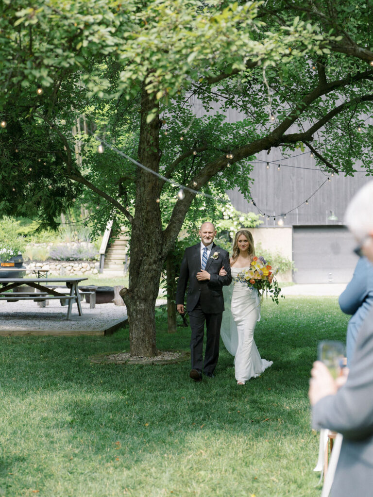 Bride walking down the aisle with her father under a canopy of trees.
