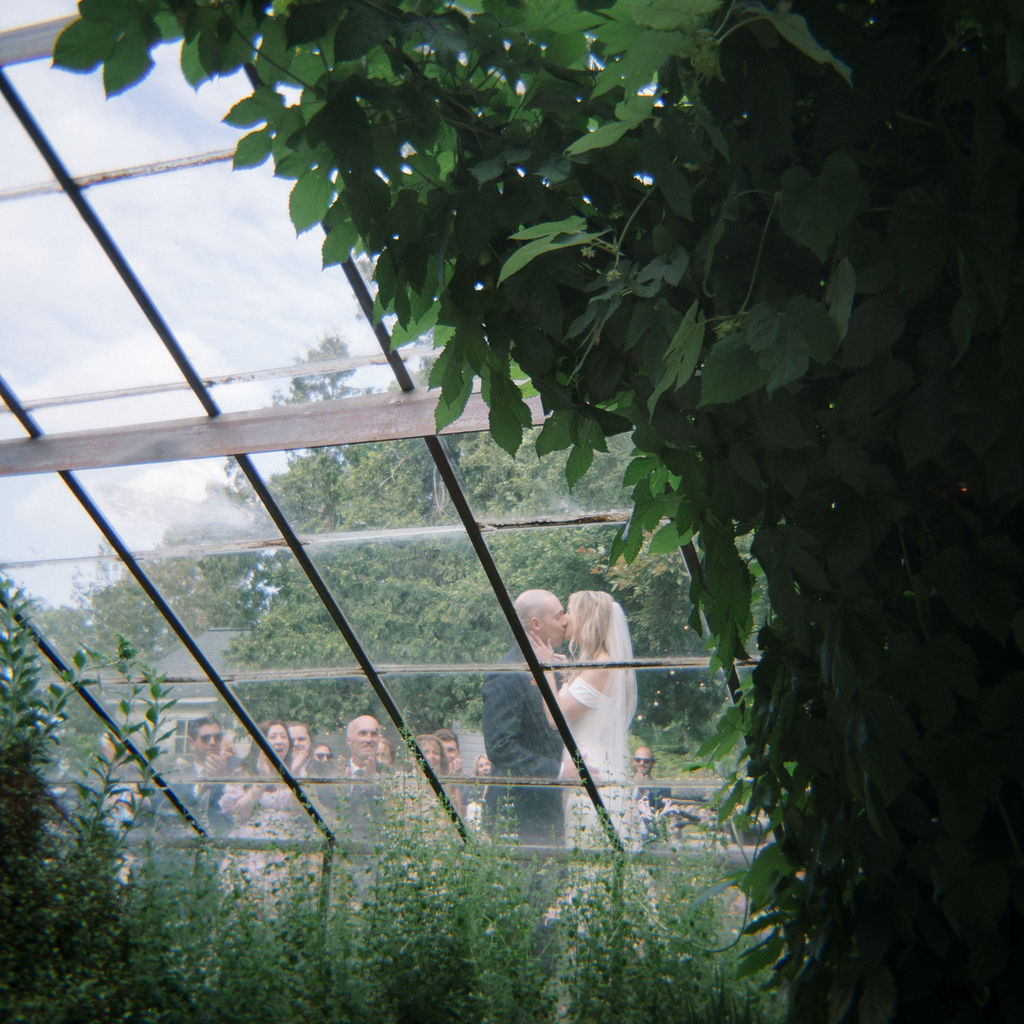 Bride and groom kissing seen through the greenhouse glass during the ceremony