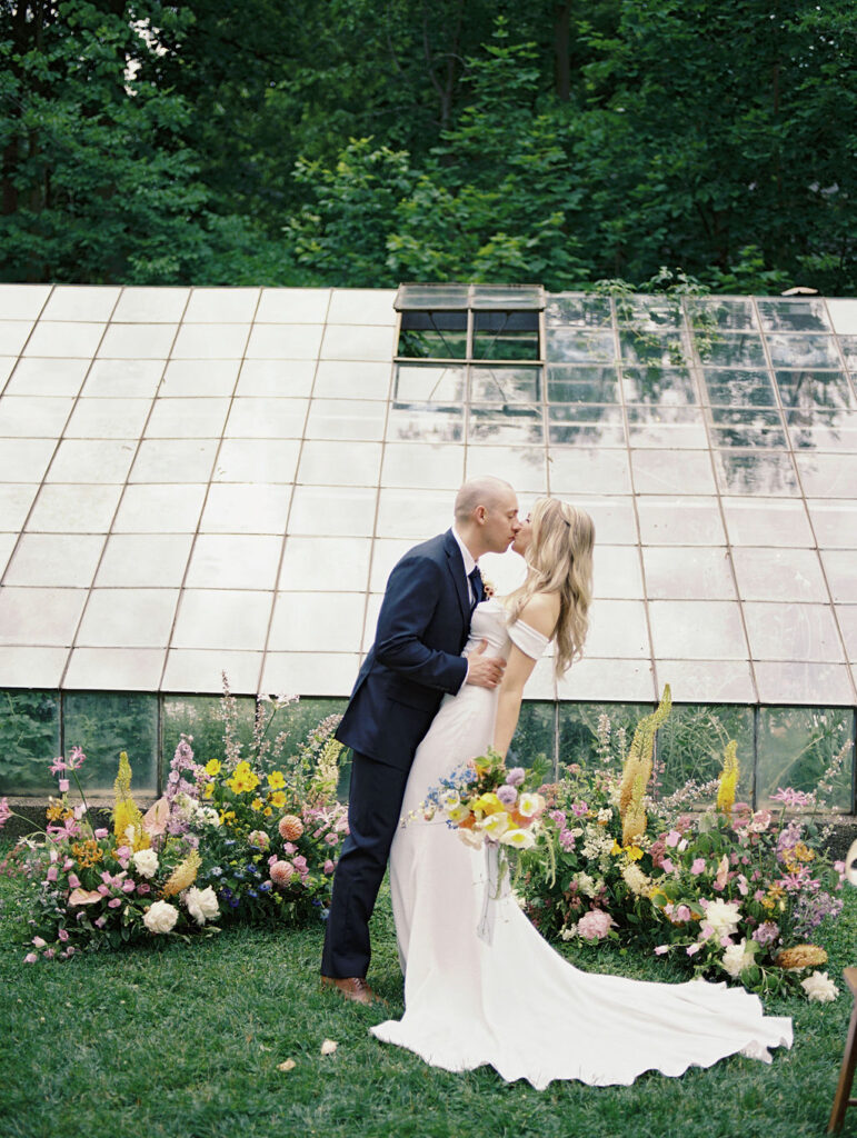 Bride and groom kissing for their Glass House Community wedding portraits in front of the greenhouse