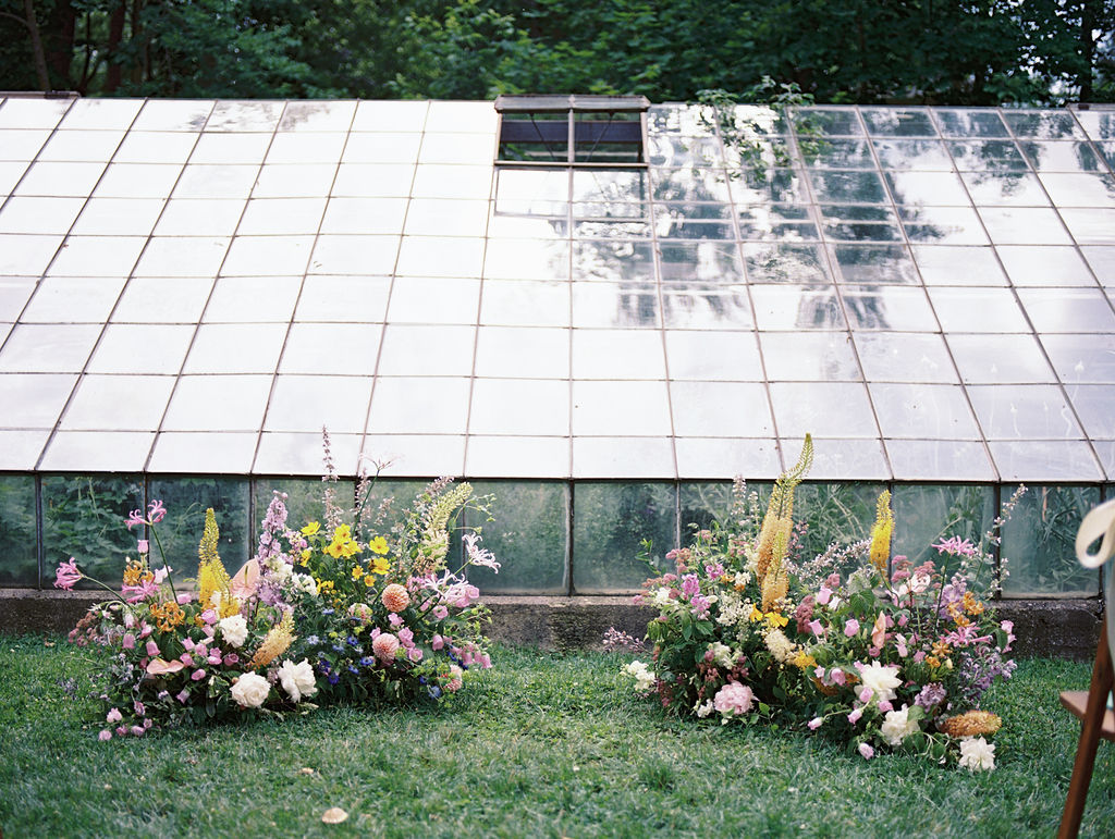 Ceremony setup with vibrant flowers and glass greenhouse backdrop.