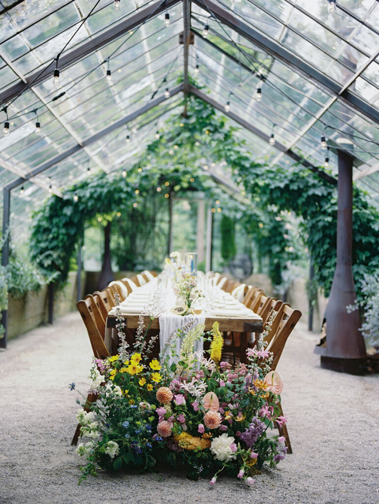 Reception table set inside The Glass House Community greenhouse with colorful florals and string lights.