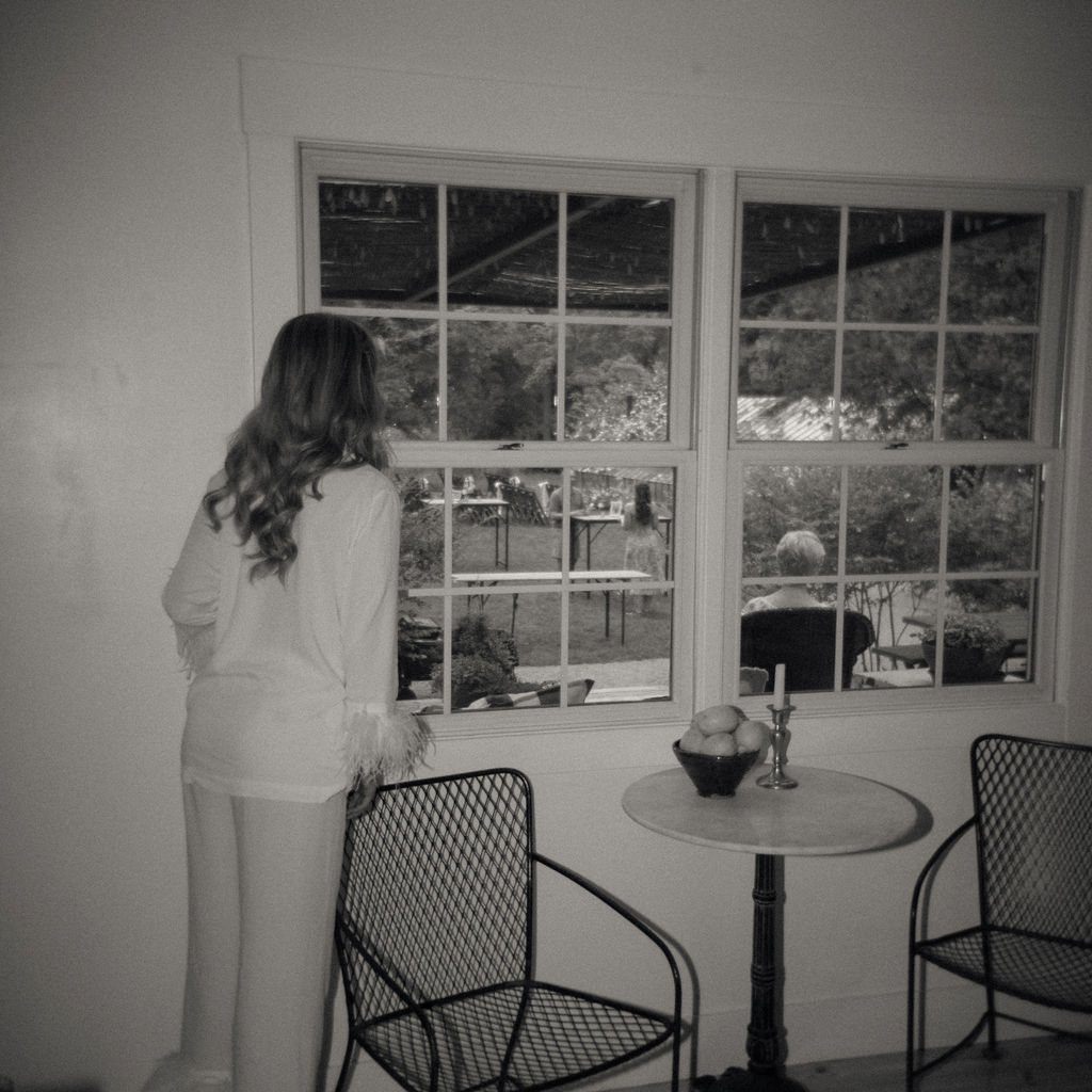 Bride in feather-trimmed pajamas looking out the window before her wedding.