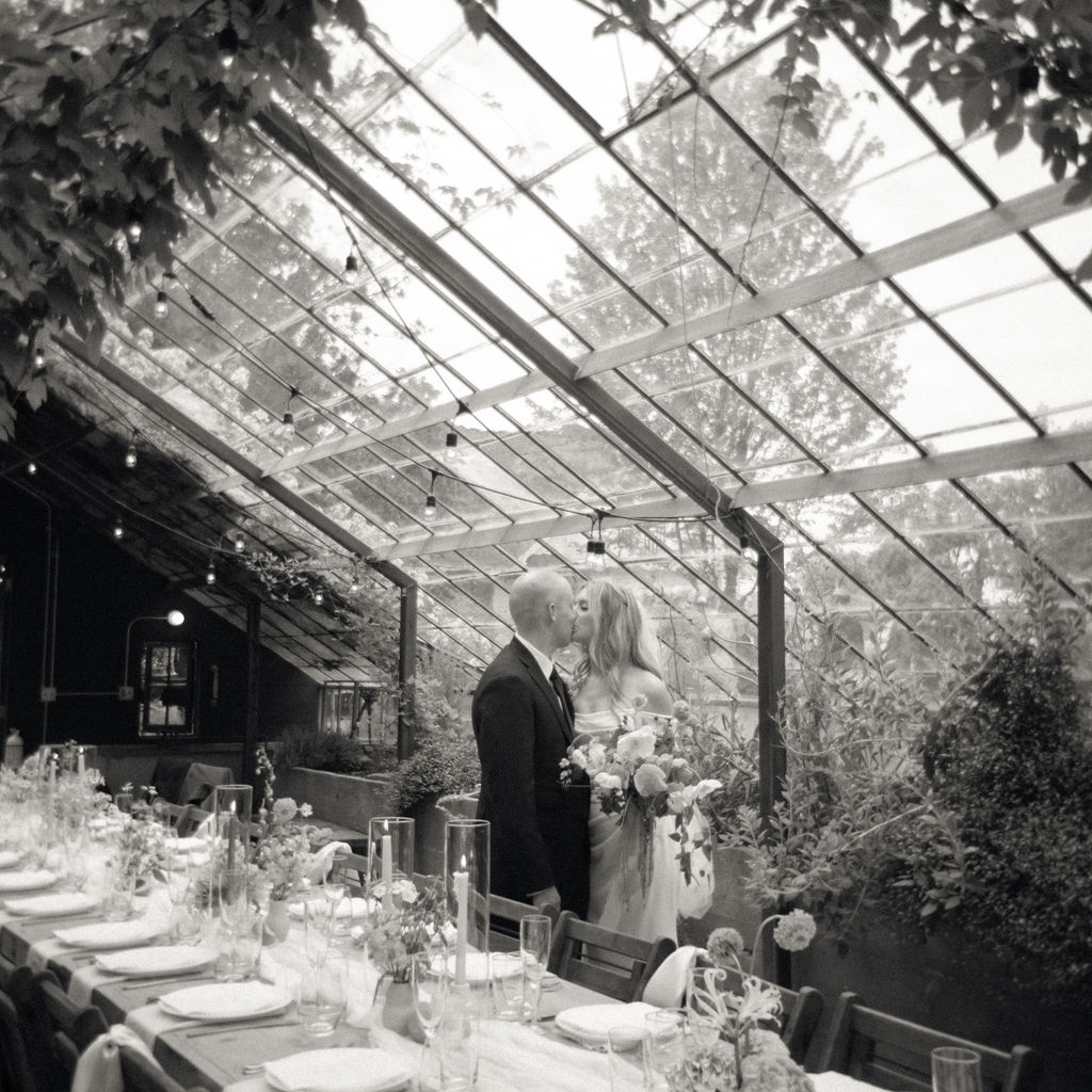 Black and white photo of a bride and groom kissing in the greenhouse at Glass House Community