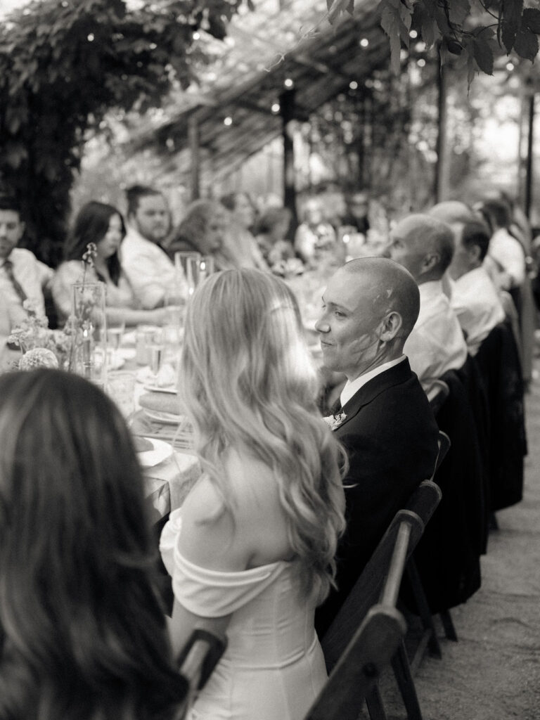 Candid black and white photo of a groom and bride smiling at each other during their Glass House Community wedding reception in the greenhouse