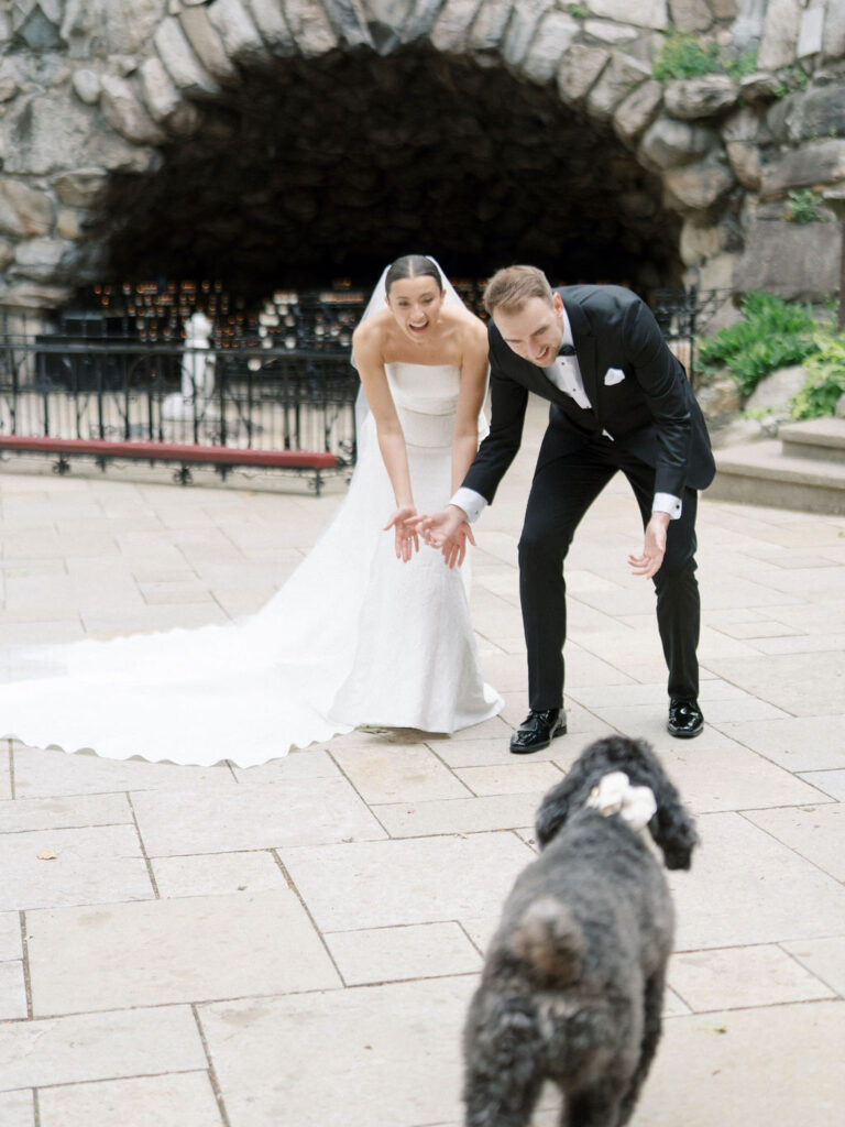Bride and groom crouching down to greet their dog wearing a bowtie at the Grotto during Saint Mary’s Notre Dame wedding – Cynthia Mae Photo.