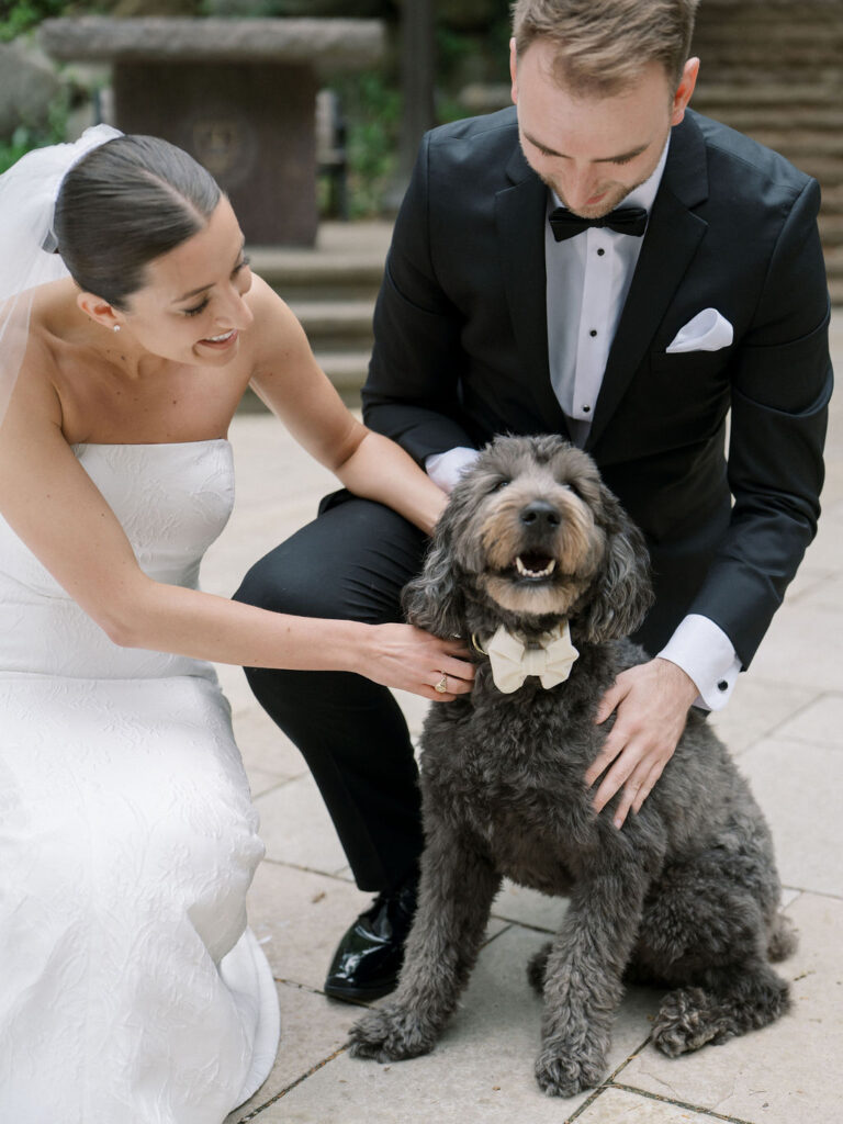 Bride and groom crouching down to greet their dog wearing a bowtie at the Grotto during Saint Mary’s Notre Dame wedding – Cynthia Mae Photo.