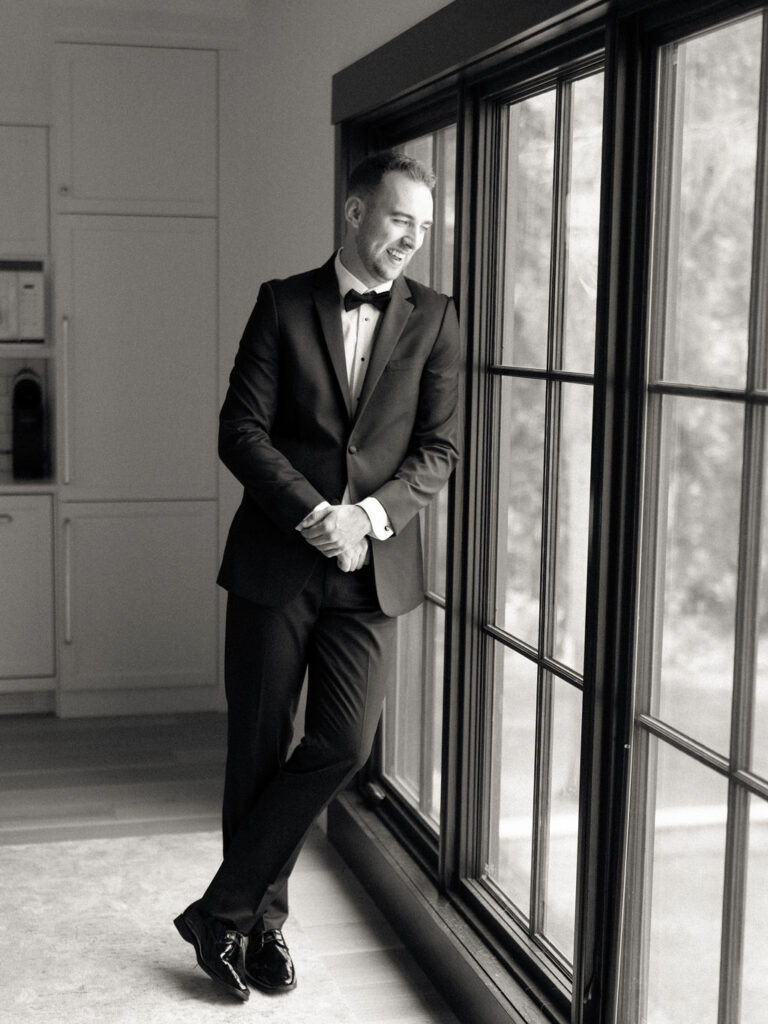 Black and white indoor grooms portrait of the groom laughing while looking out a window