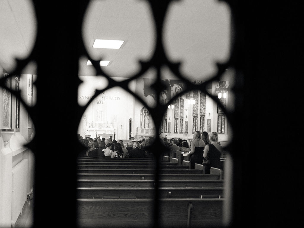Black and white photo taken through decorative window showing guests seated during Church of Our Lady of Loretto ceremony – Cynthia Mae Photo.