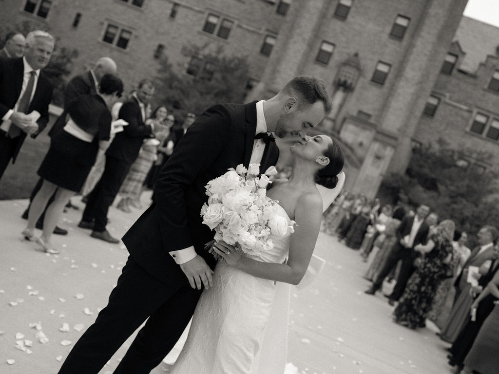 Black and white photo of a bride and groom kissing during petal toss exit outside Church of Our Lady of Loretto at Saint Mary’s College – Cynthia Mae Photo.