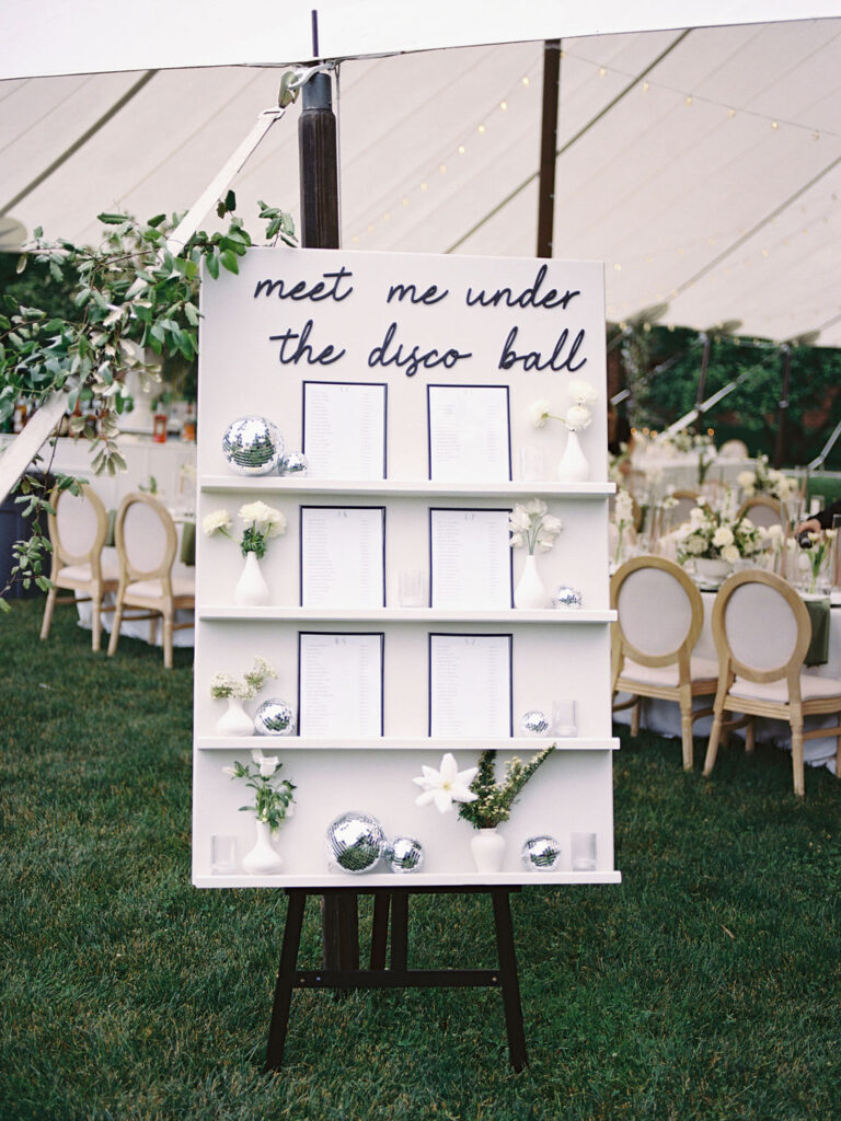 Reception seating chart with “Meet Me Under the Disco Ball” sign surrounded by disco balls and greenery at Saint Mary’s College tented wedding – Cynthia Mae Photo.