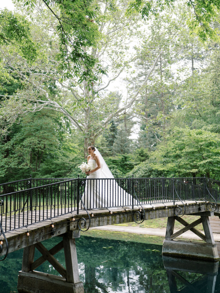 Bride walking across bridge surrounded by greenery for first look with bridesmaids at Saint Mary’s Notre Dame wedding – Cynthia Mae Photo.