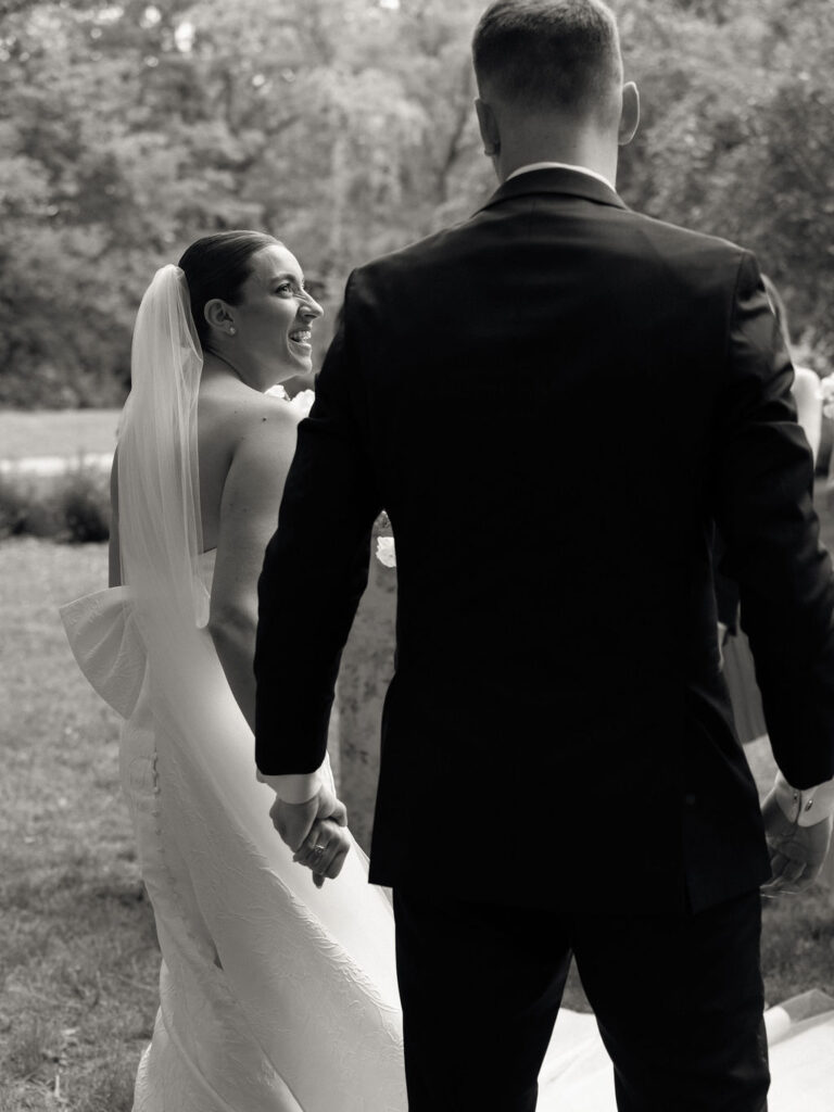 Candid black and white photo of a bride and groom holding hands and walking outdoors with their wedding party
