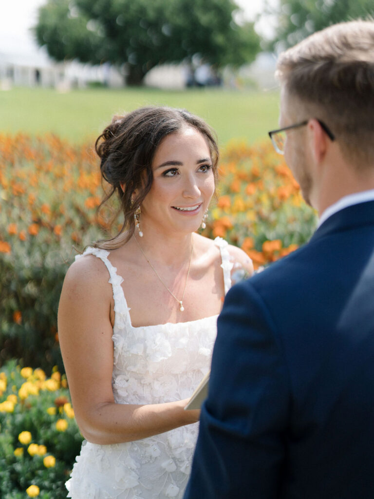 Close up shot of the bride smiling at the groom as she reads her vows to him in the gardens at Of The Land wedding venue in Marshall, Michigan