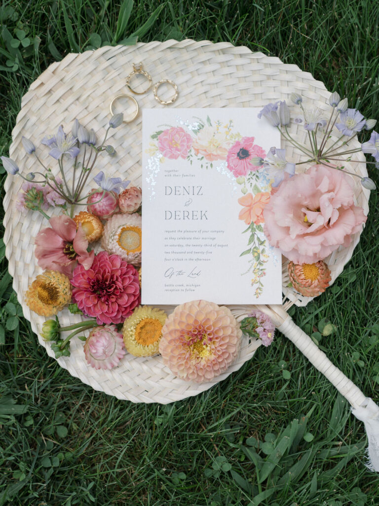Floral flatlay with wedding rings and invitation on woven fan, photographed at an Of The Land wedding.