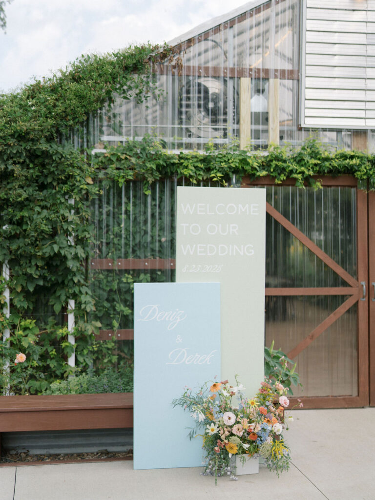 Wedding welcome signage with pastel panels and florals outside the greenhouse.
