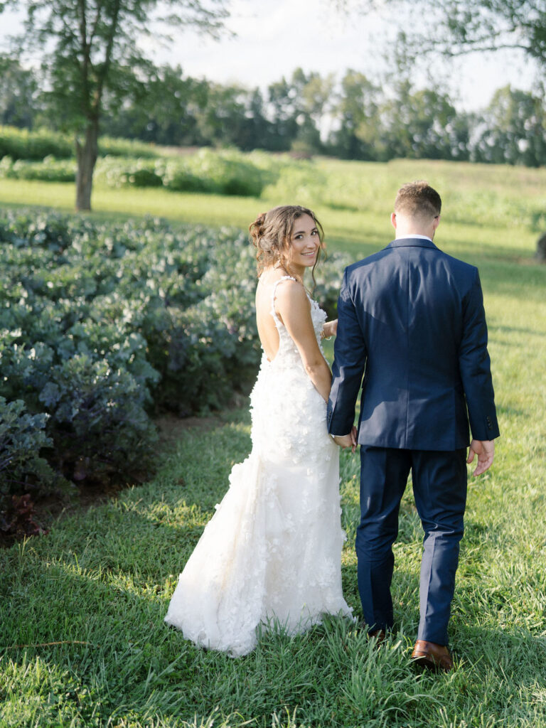 Bride looking back while walking through garden rows with groom during golden hour.