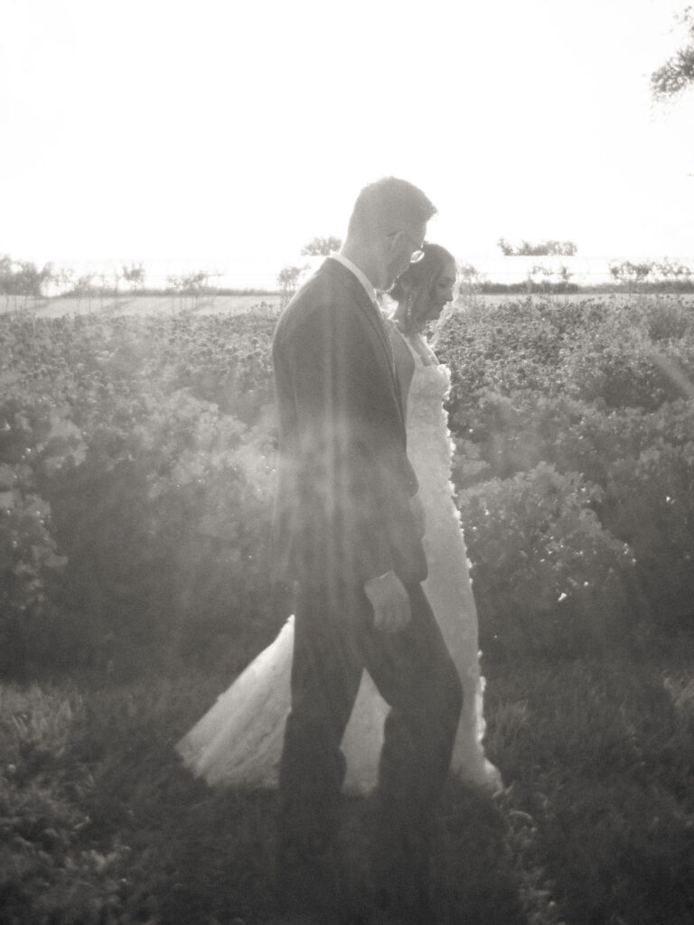 Film-style black and white photo of couple walking through sunlit fields.