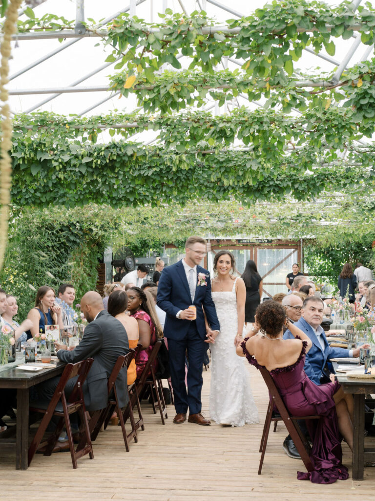 Bride and groom walk hand-in-hand through the greenhouse reception space at their Of The Land wedding.