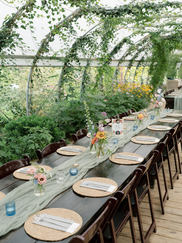 Close-up of reception tables at an Of The Land wedding, featuring woven chargers, soft green runners, and wildflower centerpieces beneath a greenhouse canopy.