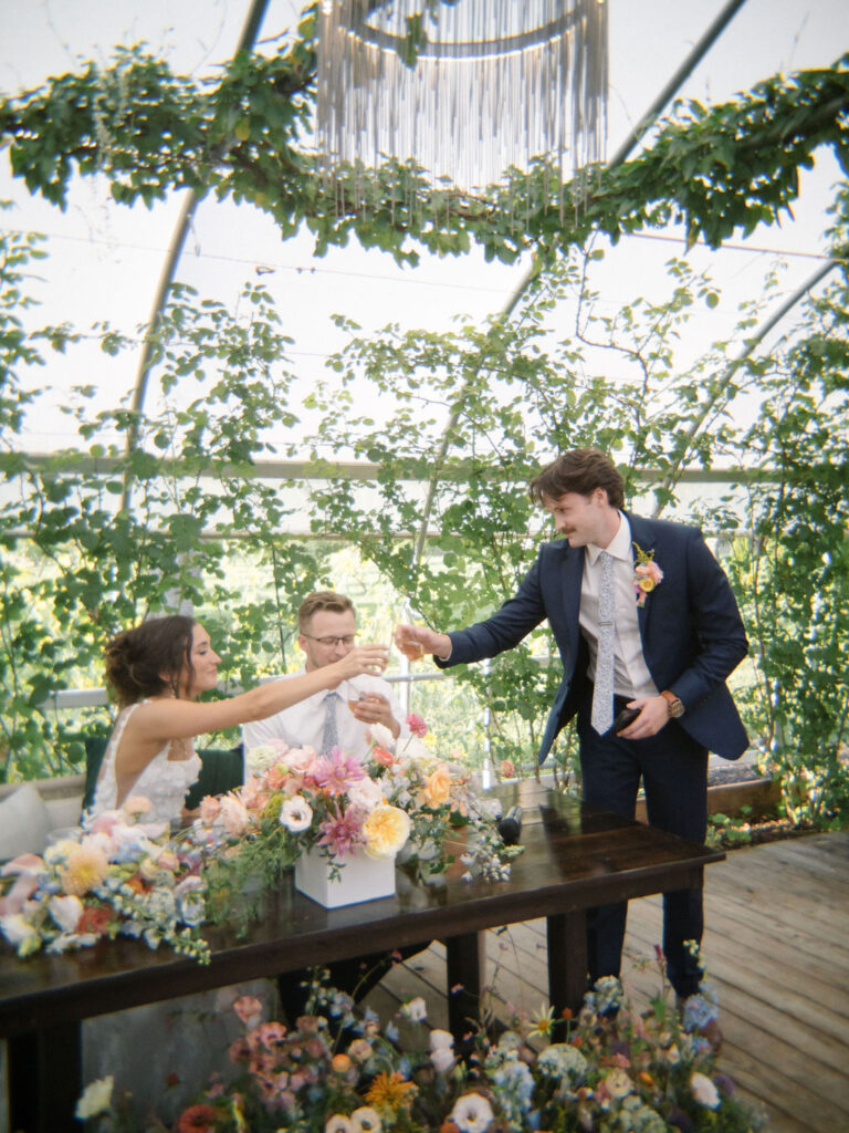 Bride, groom, and best man share a toast at their sweetheart table overflowing with seasonal florals during an Of The Land wedding reception.
