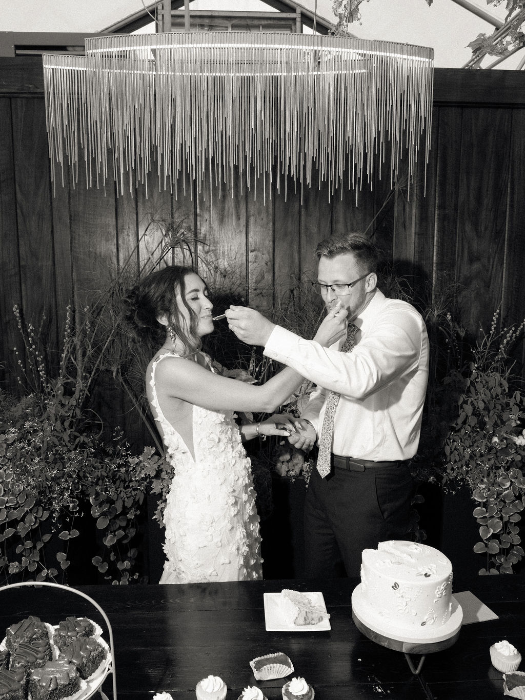 Black-and-white photo of the couple feeding each other cake beneath a modern fringe chandelier at their Of The Land wedding.