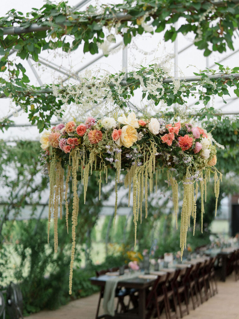 Lush floral chandelier with dahlias and trailing amaranthus hanging inside the greenhouse at an Of The Land wedding reception.