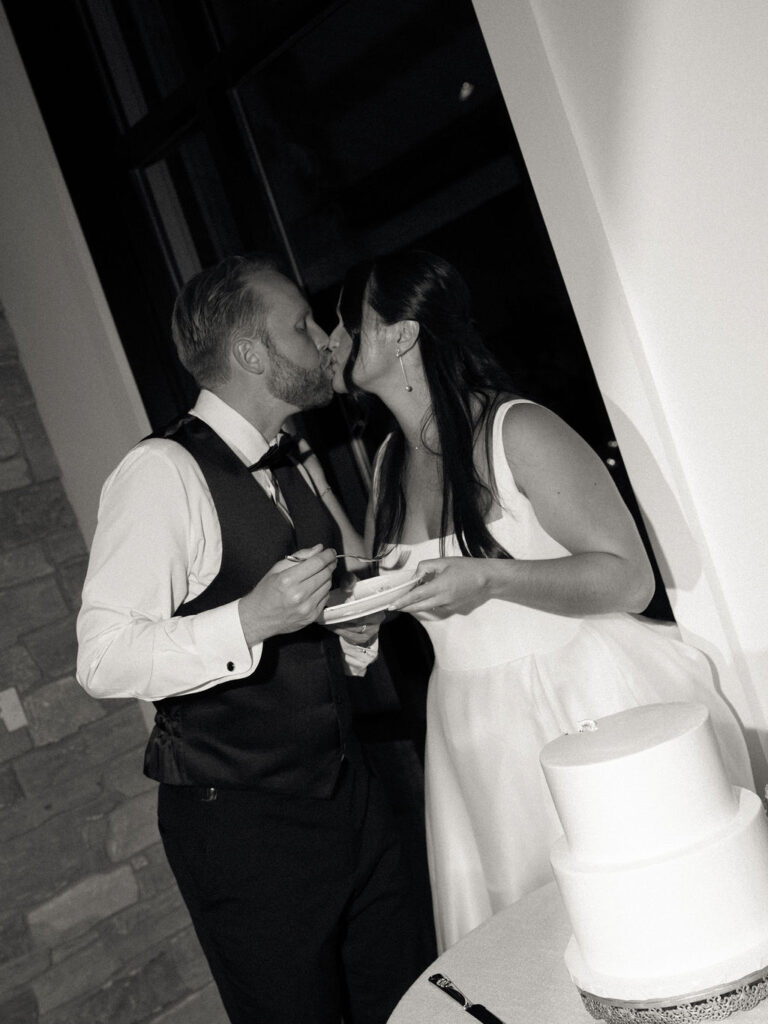 Black and white flash photo of a bride and groom kissing during their cake cutting.