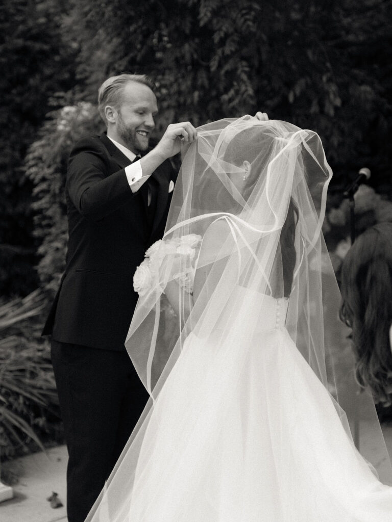 Black and white photo of the groom lifting the brides veil over her face for their Bay Pointe Woods wedding ceremony.