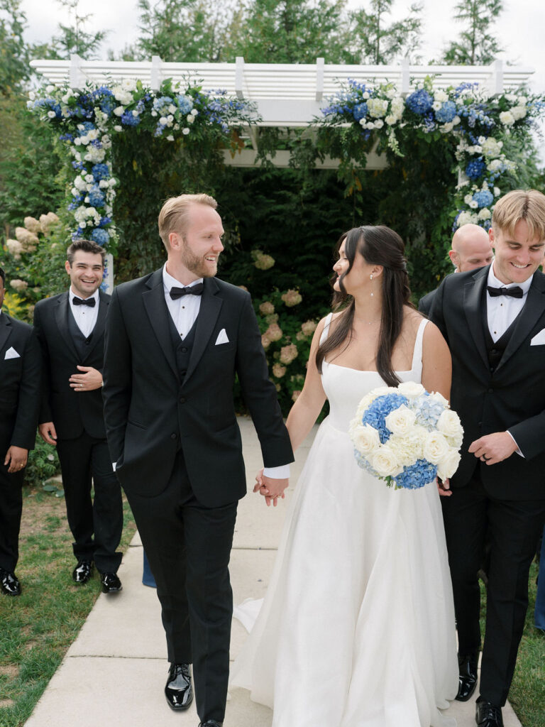 Bride and groom walking hand in hand with their wedding party near the ceremony pergola.