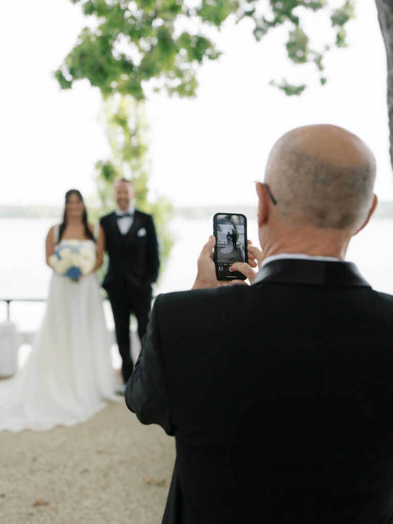 Father of the bride taking a photo on his phone of the bride and groom.