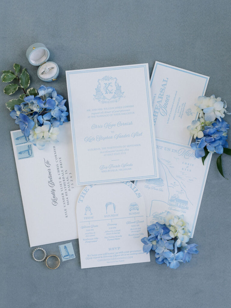 Flat lay of blue and white wedding invitations with rings and florals for a Bay Pointe Woods wedding.