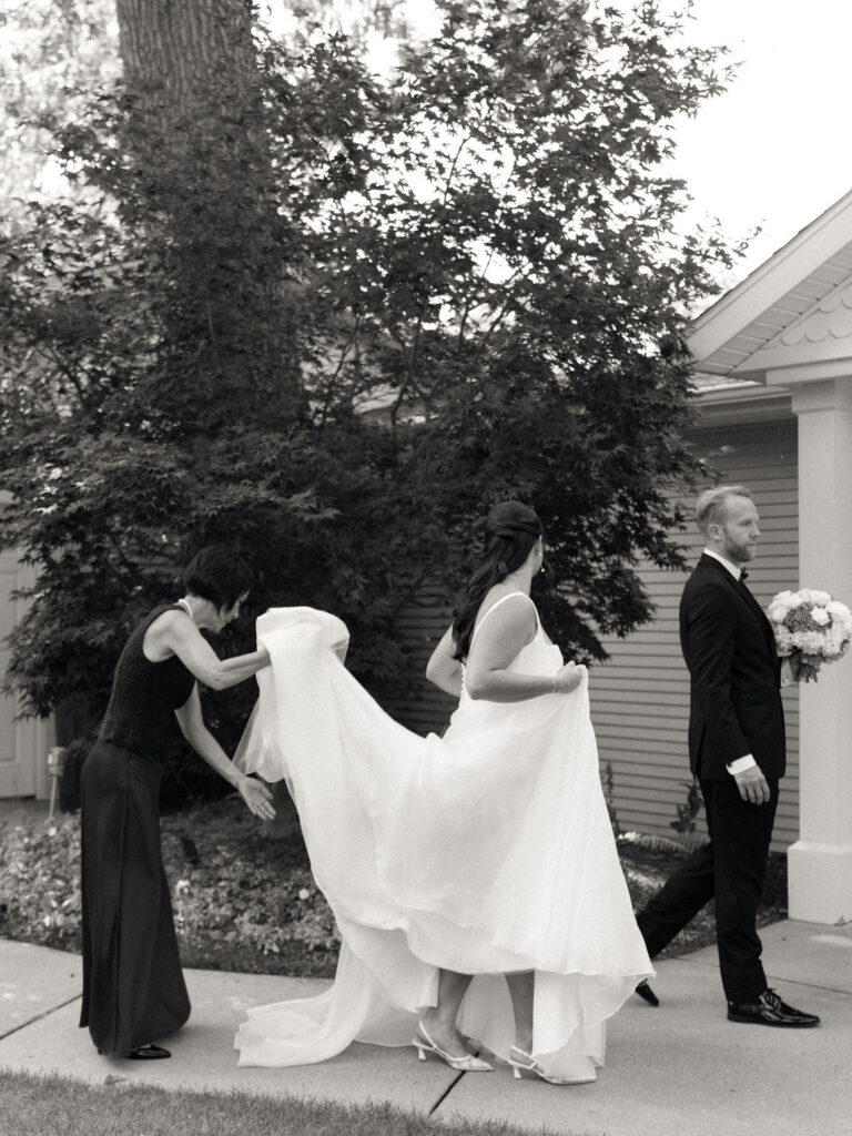 Candid black and white photo of ab ride and groom walking together outside with the brides mother holding her train.